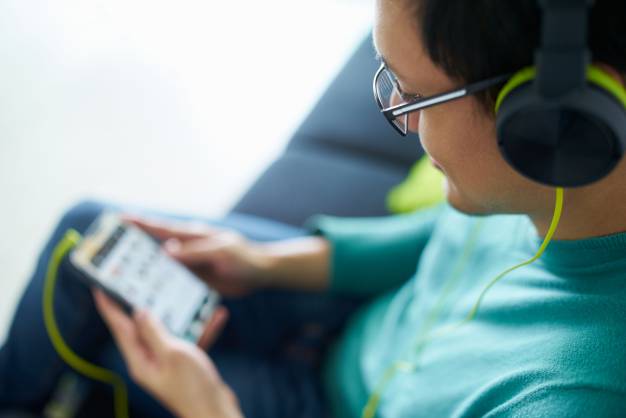 Chinese man relaxes on sofa and watches podcast on mobile phone, listening with green big earphones. Copy space